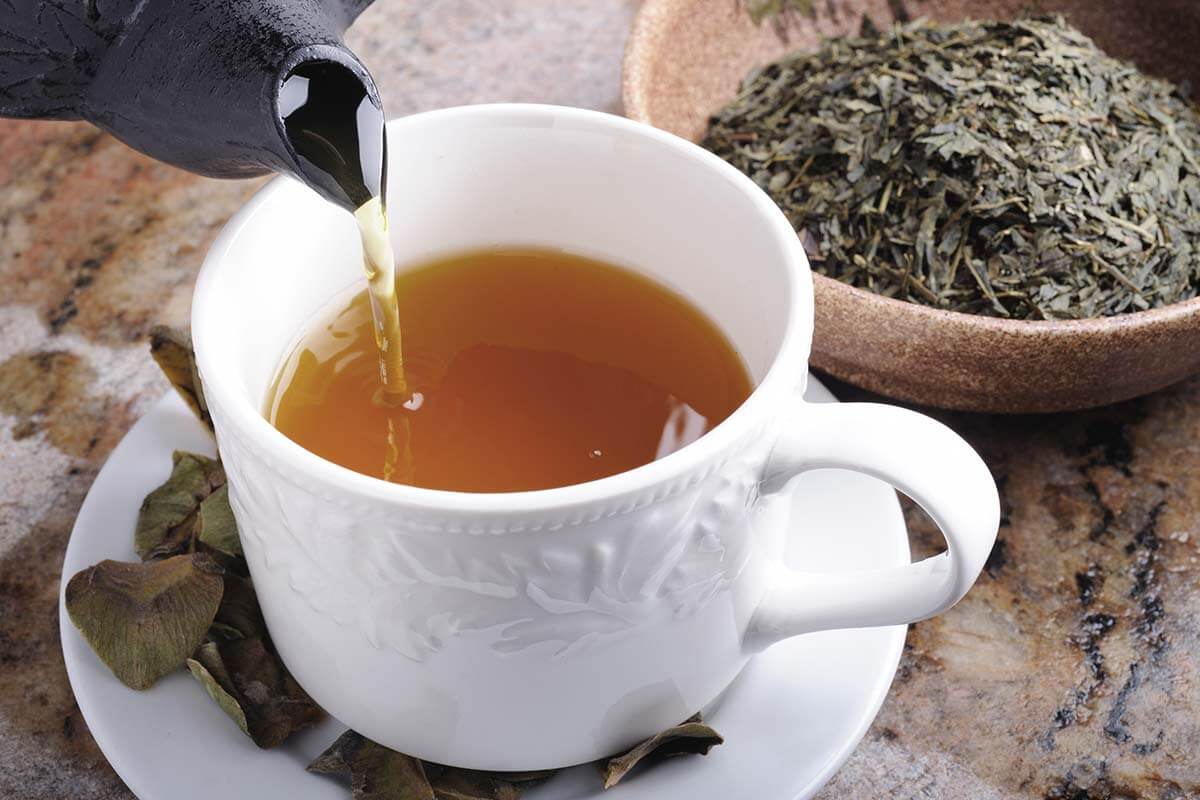 یک عدد قوری در حال ریختن چای سبز درون یک فنجان سفید به همراه یک کاسه چای سبز خشک